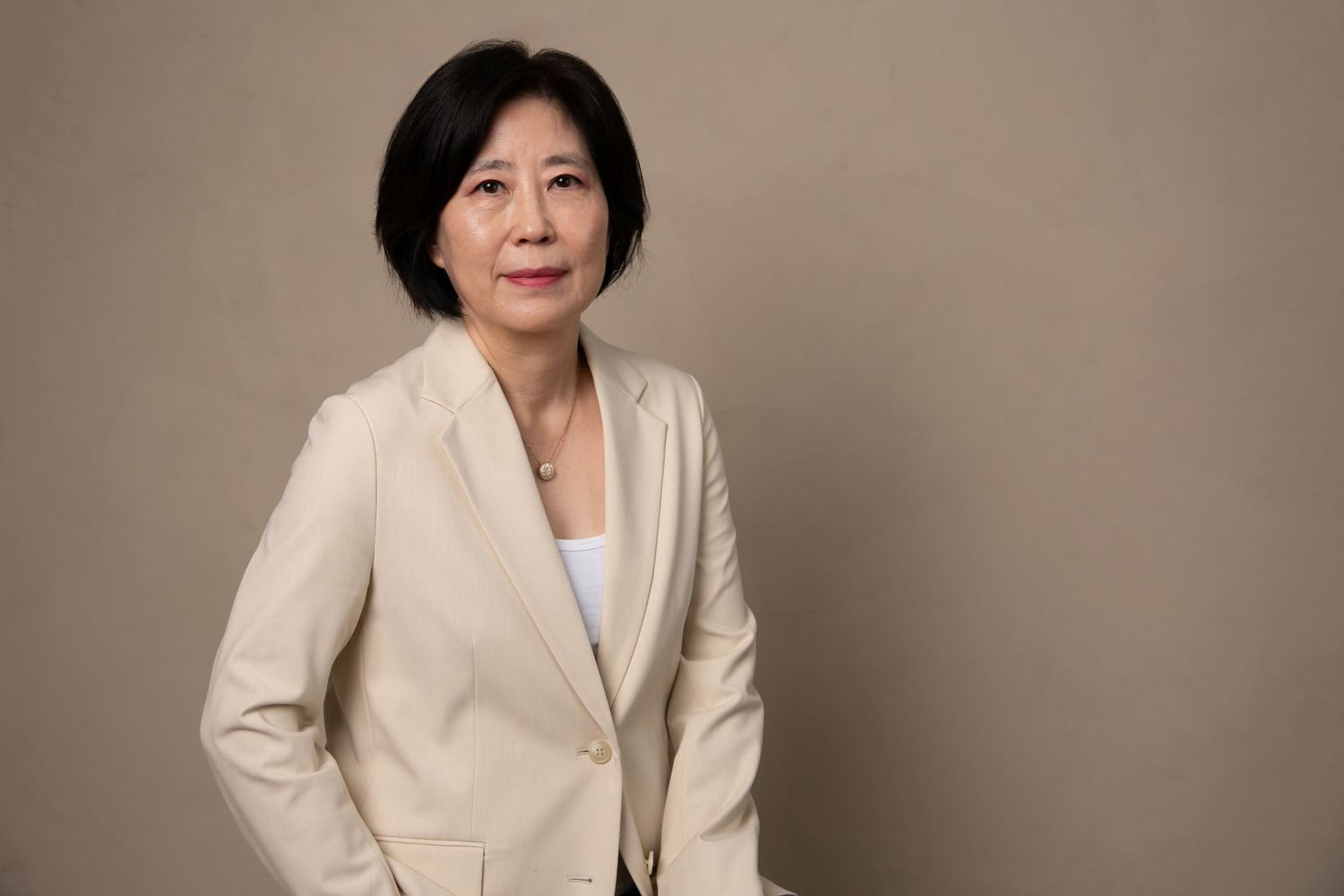 Professional Asian woman in beige blazer posing confidently against a neutral studio background.