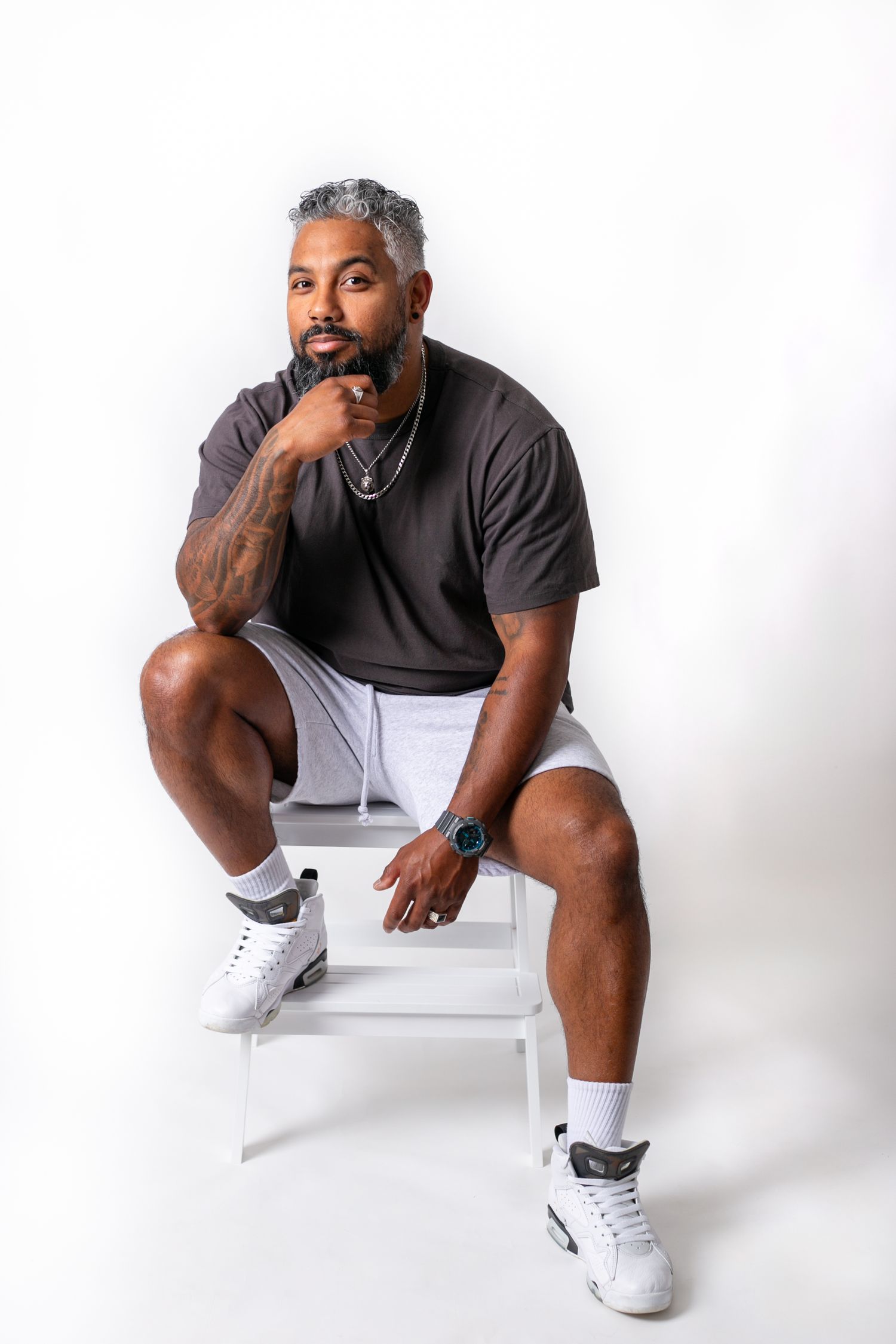 Person in black shirt and white shorts sits thoughtfully on white chair in studio setting.