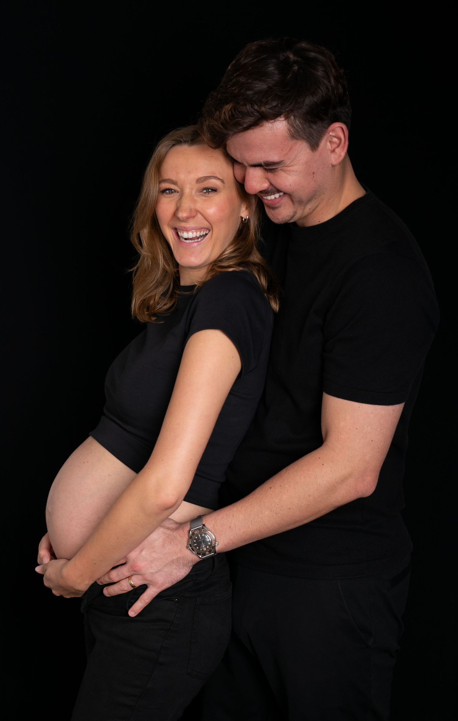 A couple wearing black clothing embraces against a dark background in a series of intimate maternity photos.