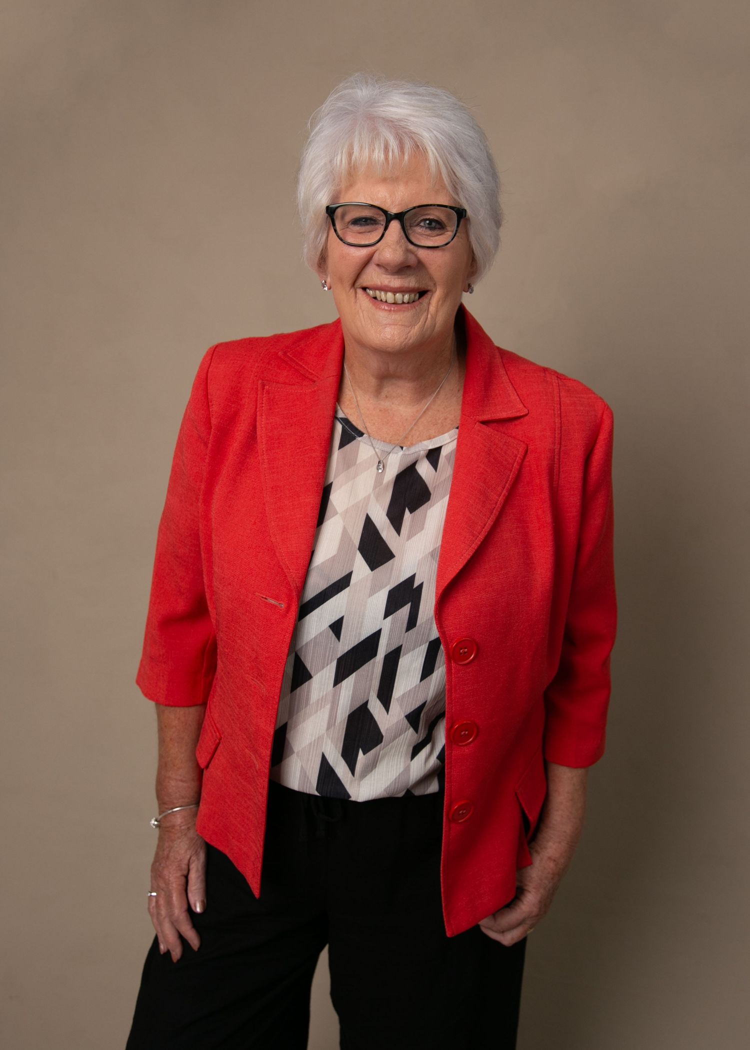 Smiling senior woman with white hair and glasses wearing a red blazer and patterned blouse, standing confidently.