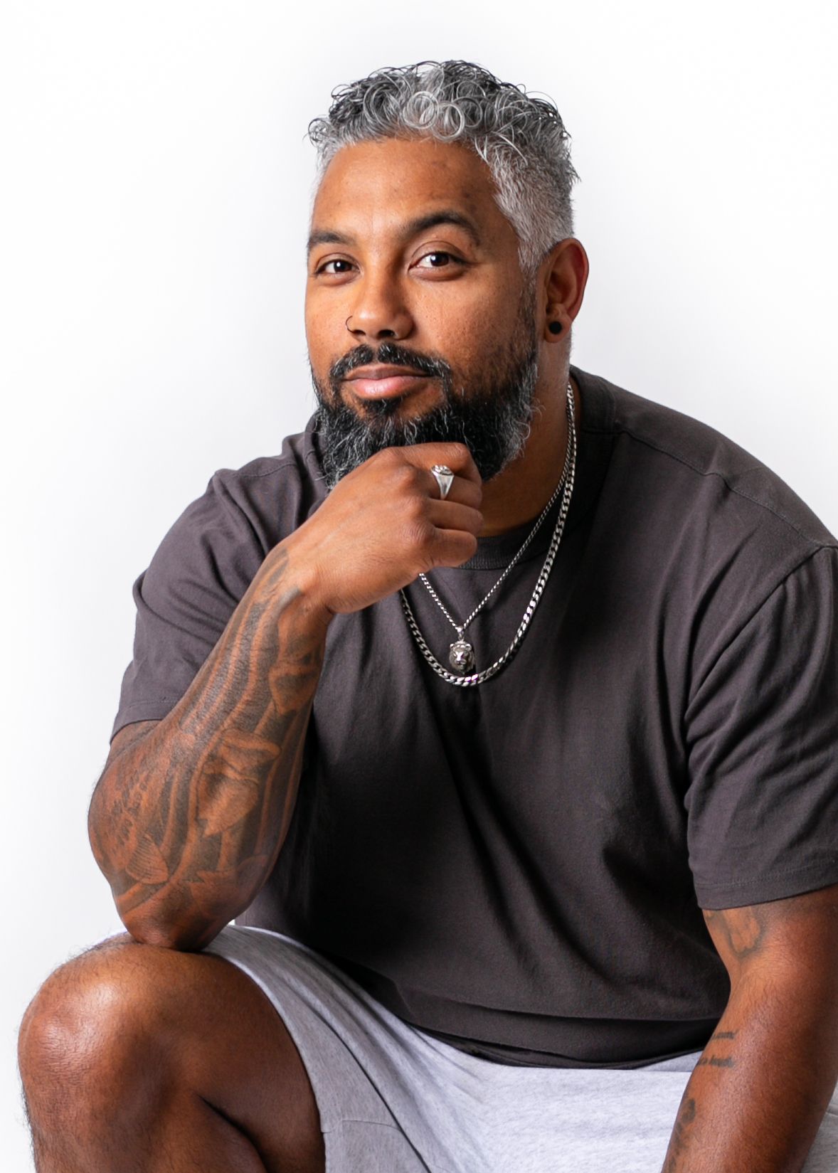 Muscular man with gray-streaked hair and beard, tattoos, wearing black tee and gray shorts, posing thoughtfully on white background.
