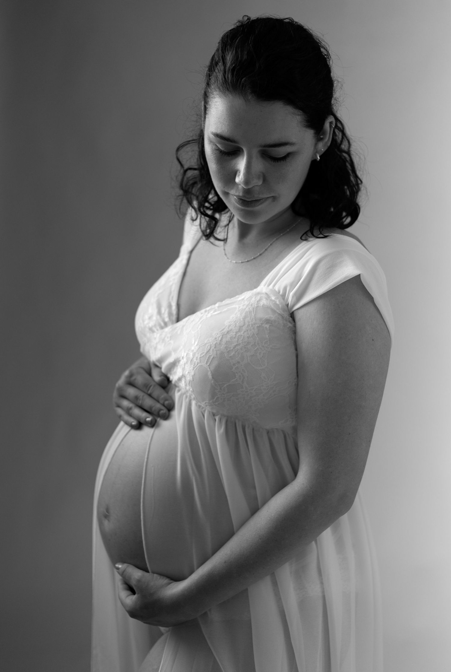 A pregnant woman in a white dress gently cradles her belly in an intimate black and white maternity portrait.