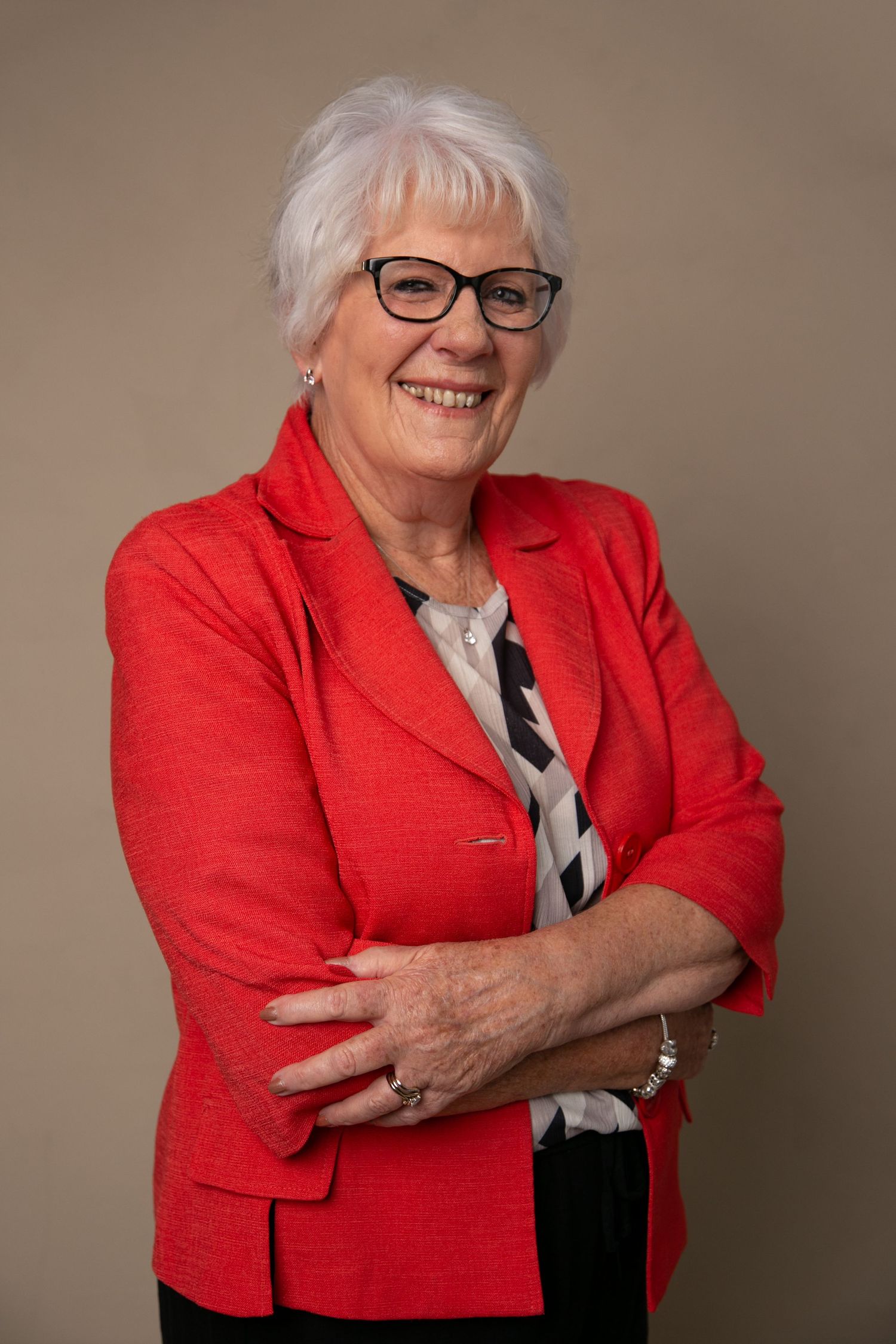 Senior woman with white hair and glasses in a red blazer, smiling with arms crossed in a professional portrait.