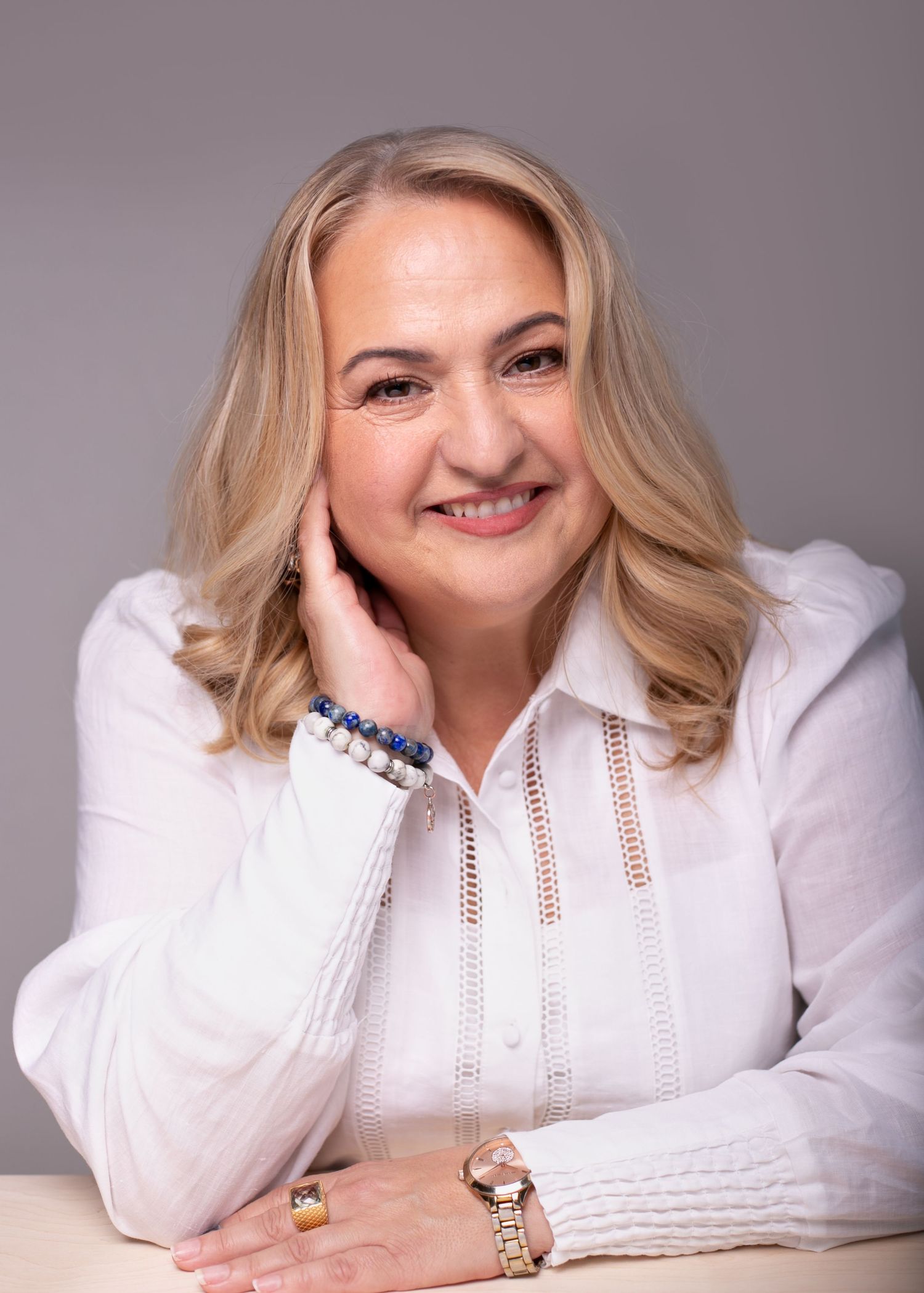 Smiling middle-aged blonde woman in white blouse, resting chin on hand, wearing bracelet and watch, professional portrait.