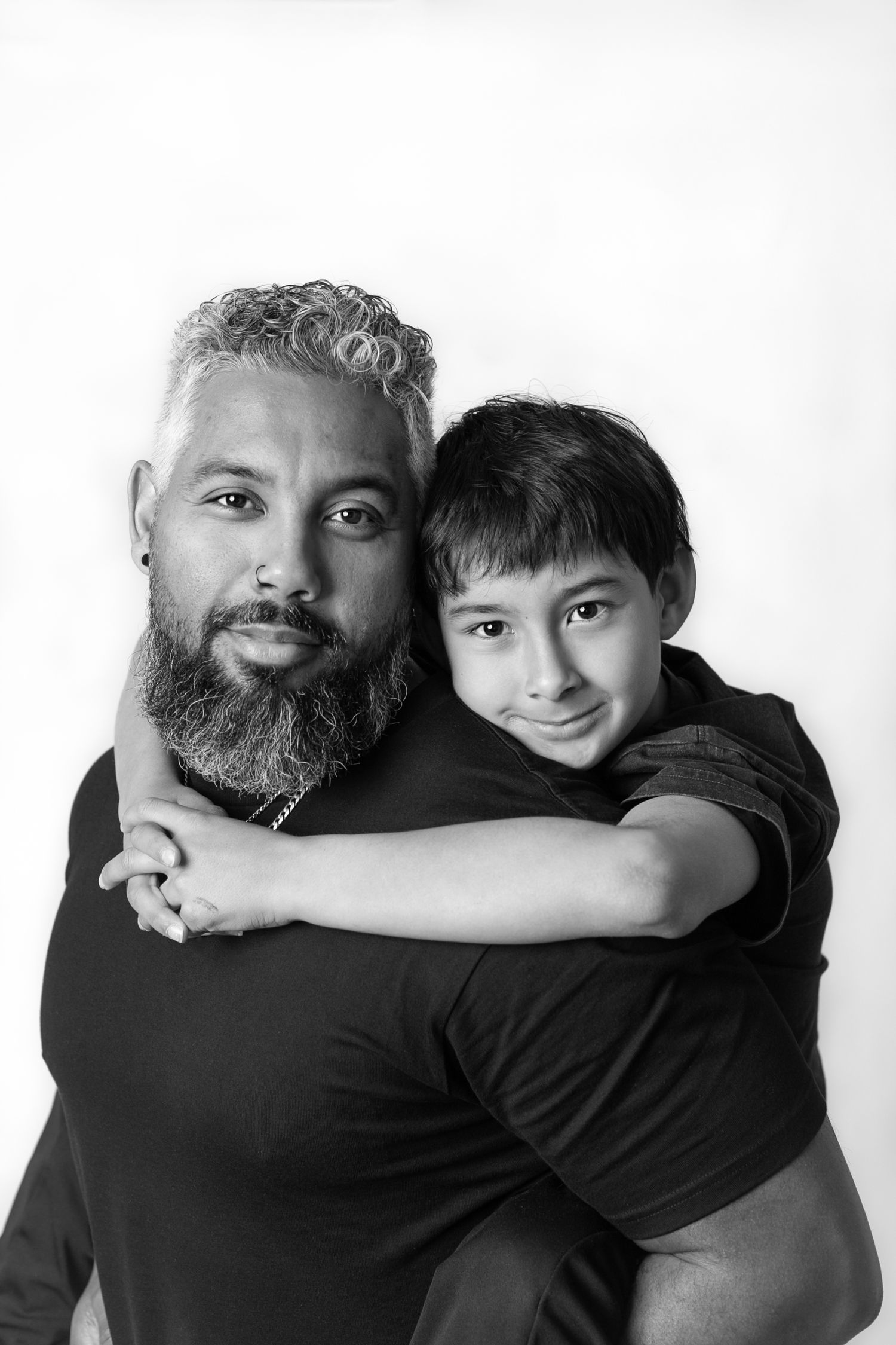 Black and white portrait of a bearded man with a young boy hugging him from behind, both smiling warmly.