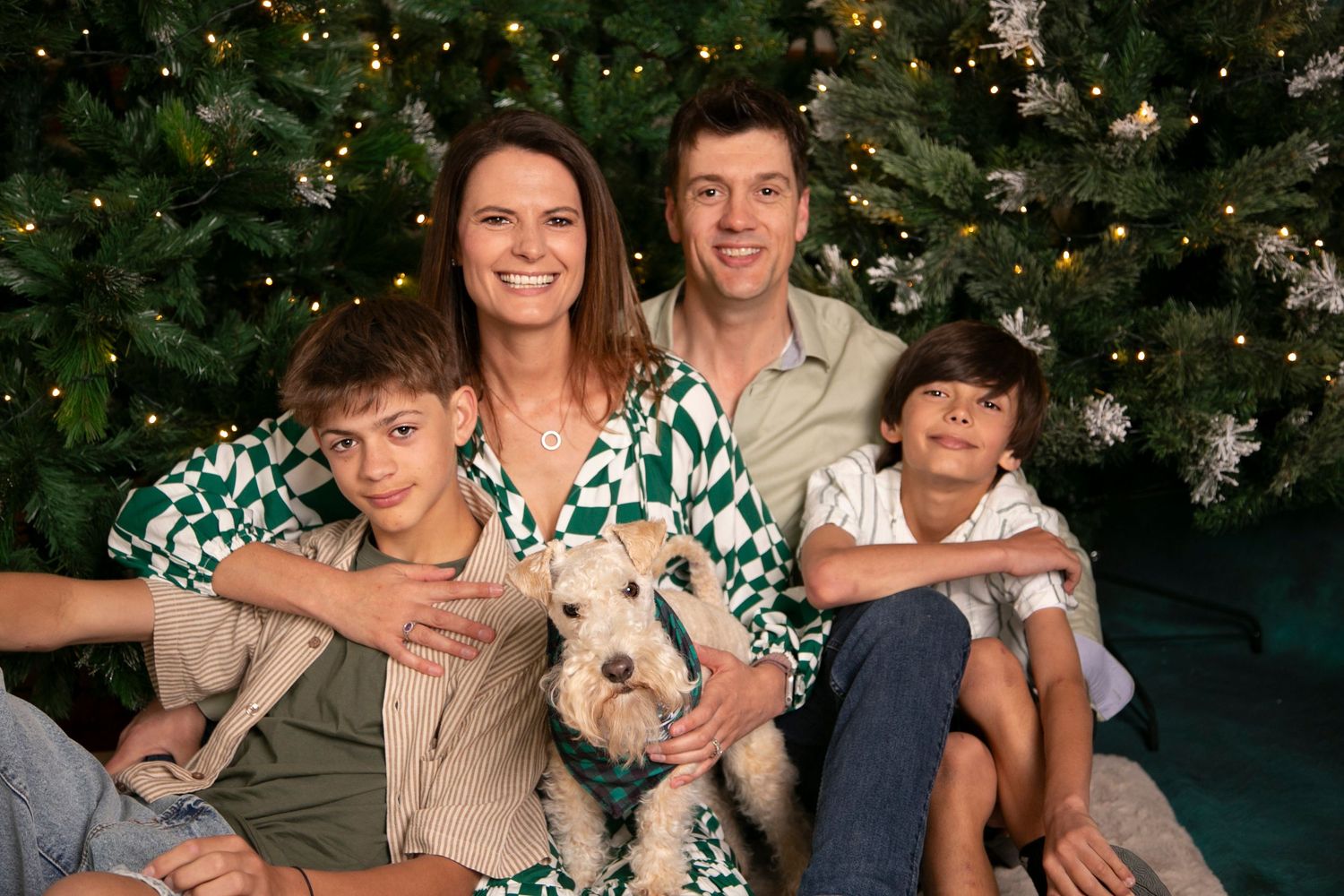 A family poses together in front of a decorated Christmas tree with their small white dog.