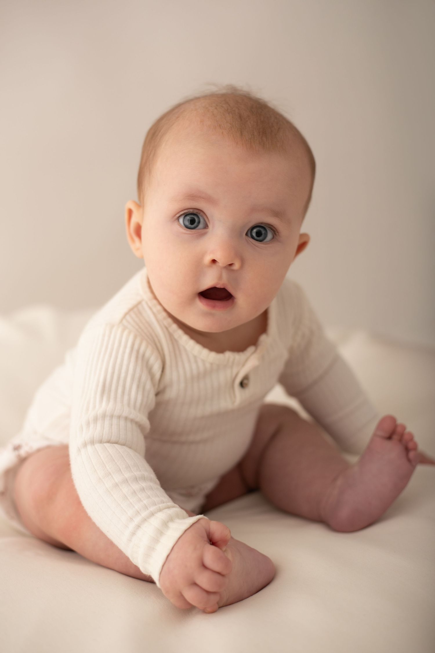 Adorable blue-eyed baby in cream ribbed onesie sitting up on white blanket with curious expression.