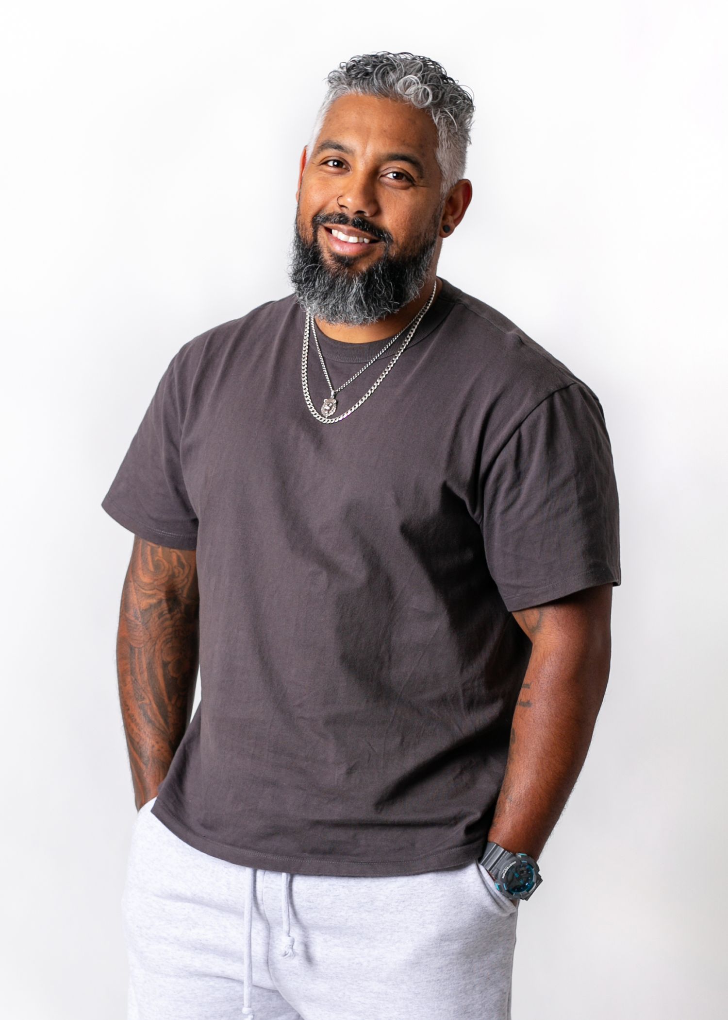 Smiling man with gray-streaked hair and beard wearing a dark gray t-shirt, chain necklaces, and light gray sweatpants.