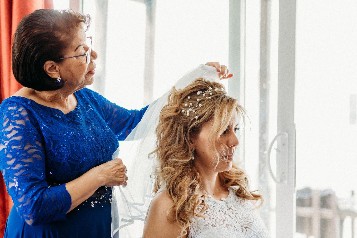Une mariée et sa mère partagent un moment de joie en se préparant près de fenêtres lumineuses le jour du  mariage.