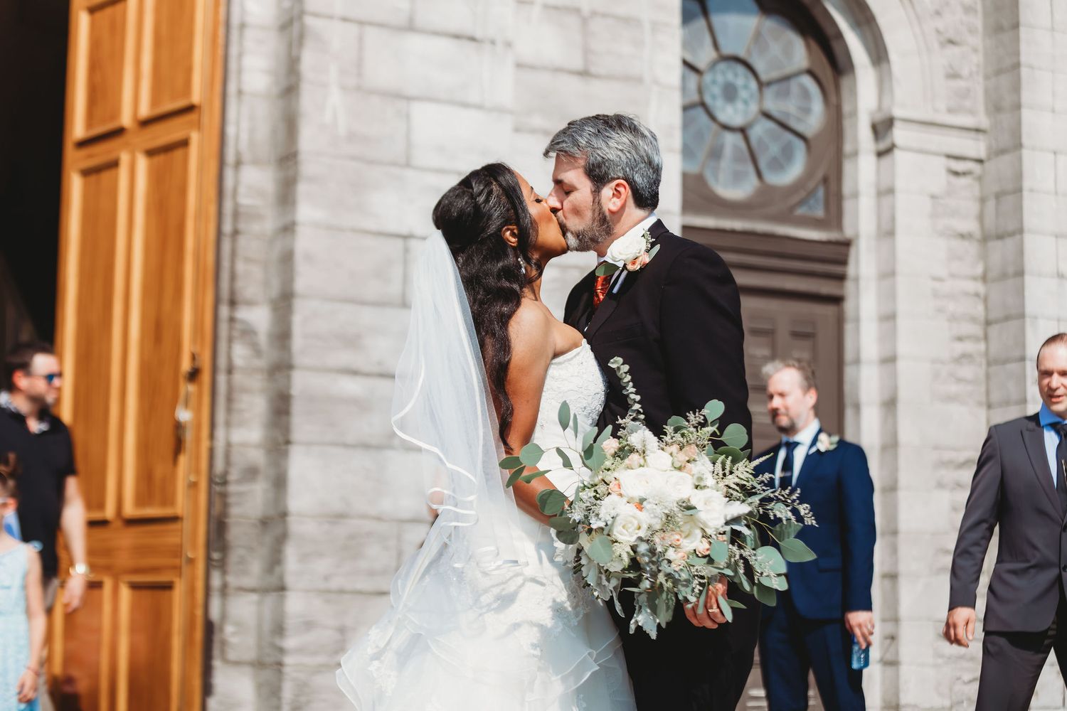 A bride and groom share a romantic kiss outside a church with wooden doors and stone walls.