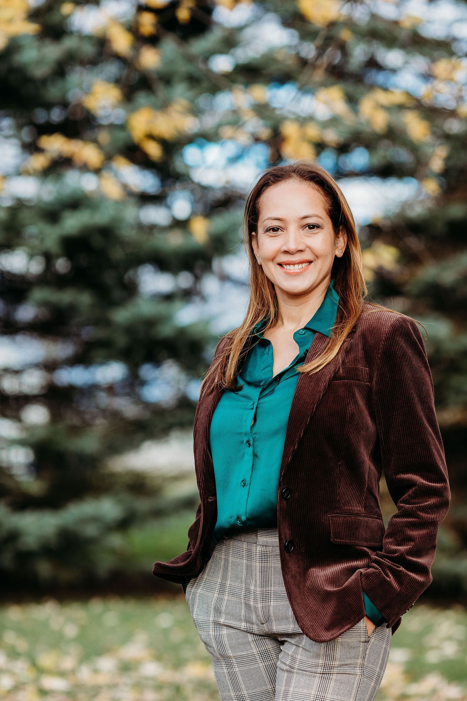 Professional portrait of someone wearing a brown velvet blazer and teal blouse against a blurred autumn background.
