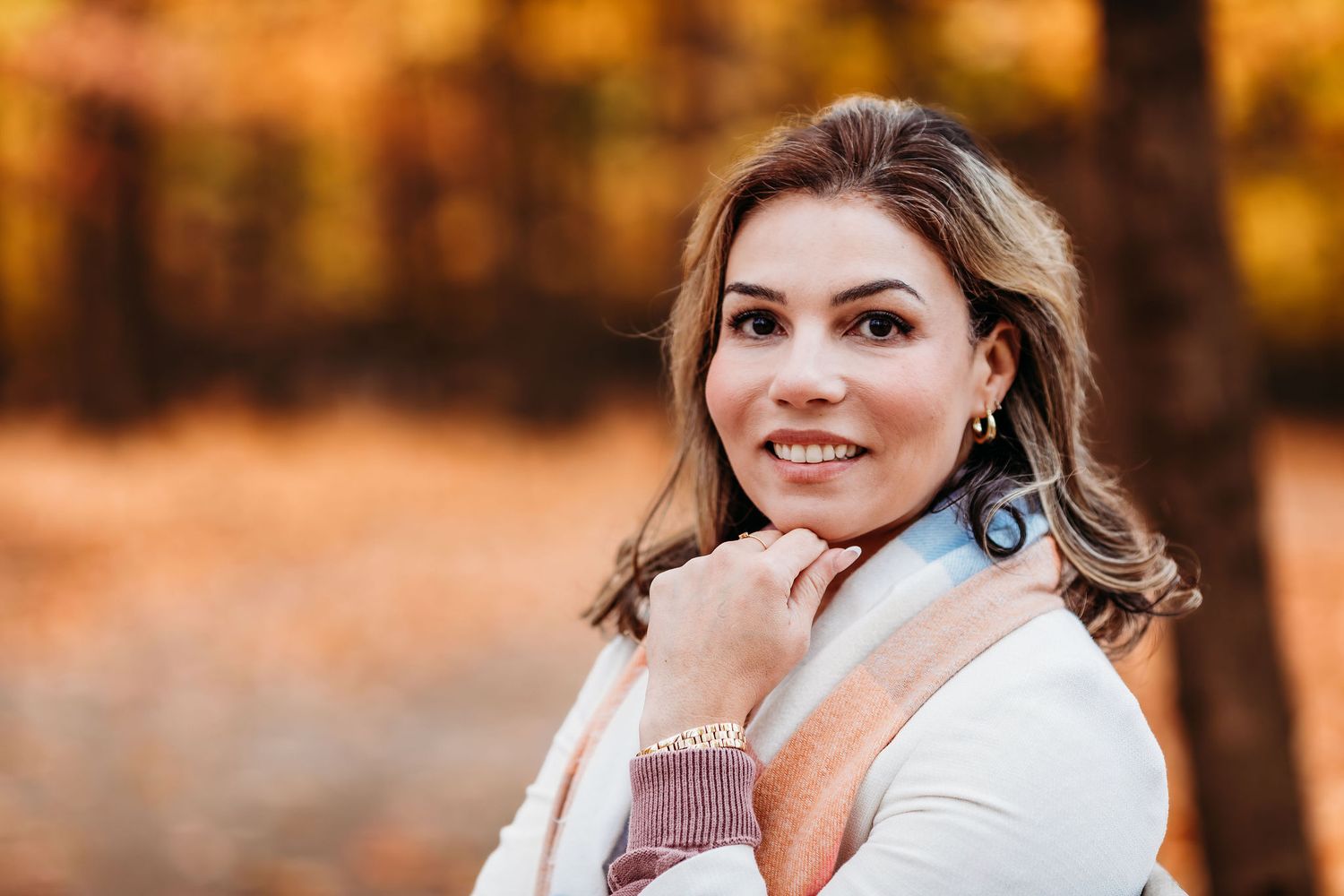Portrait of a woman smiling warmly in autumn setting with golden sunlight and wearing a white sweater and blue earrings.