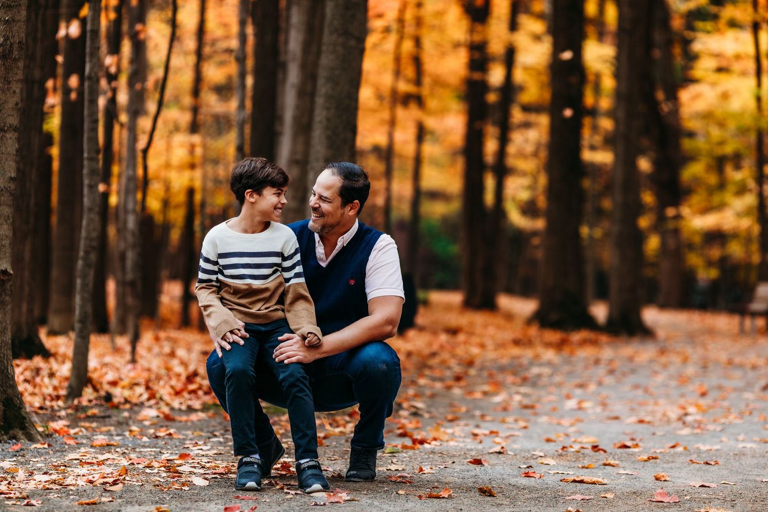 Un père et son fils assis sur des feuilles mortes d'automne dans une forêt où filtre une douce lumière dorée à travers les arbres.