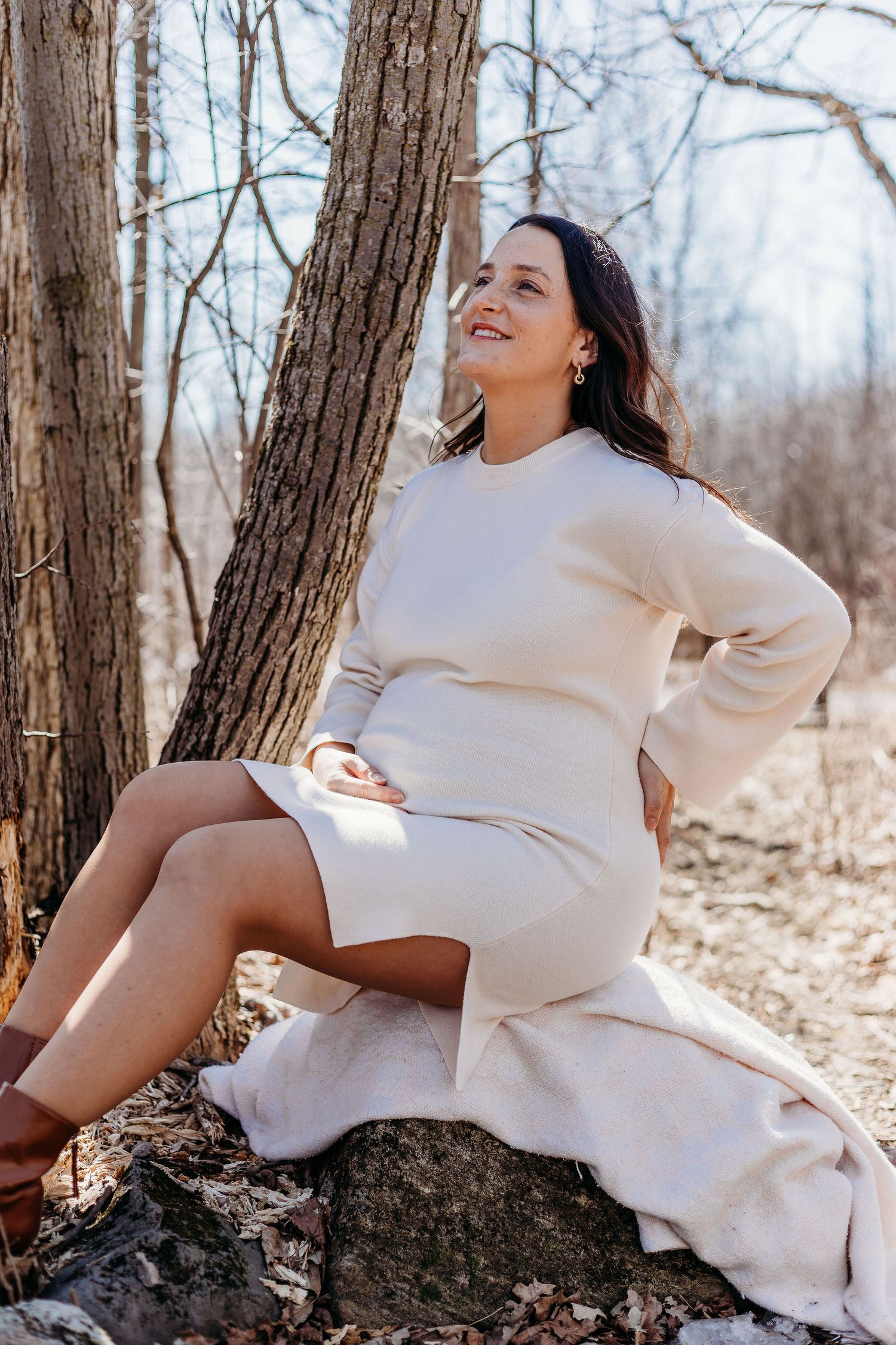 A person in a cozy cream sweater dress sits on a rock in a winter forest setting, looking up with a joyful expression.