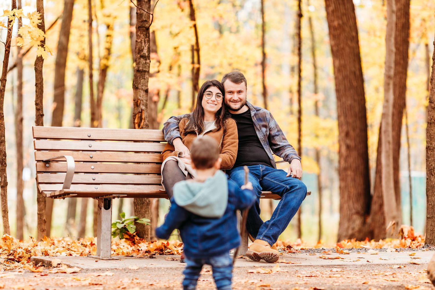 A couple sits together on a wooden bench surrounded by autumn trees with golden leaves in a peaceful forest setting.