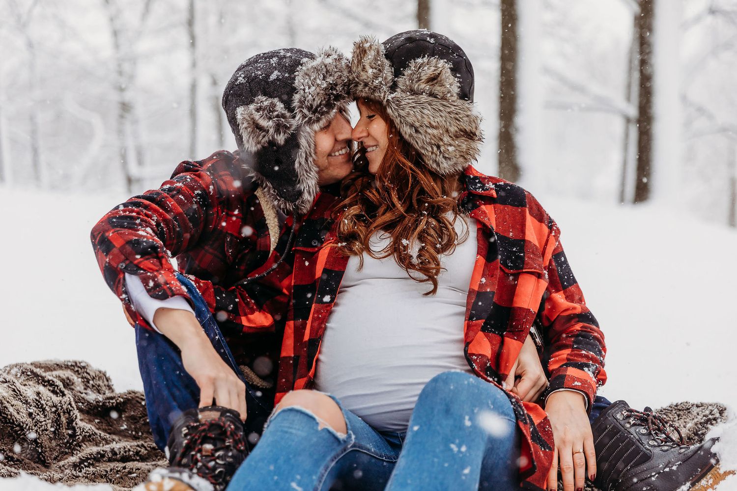 A couple in buffalo plaid and winter attire share an intimate moment in a snowy forest setting.