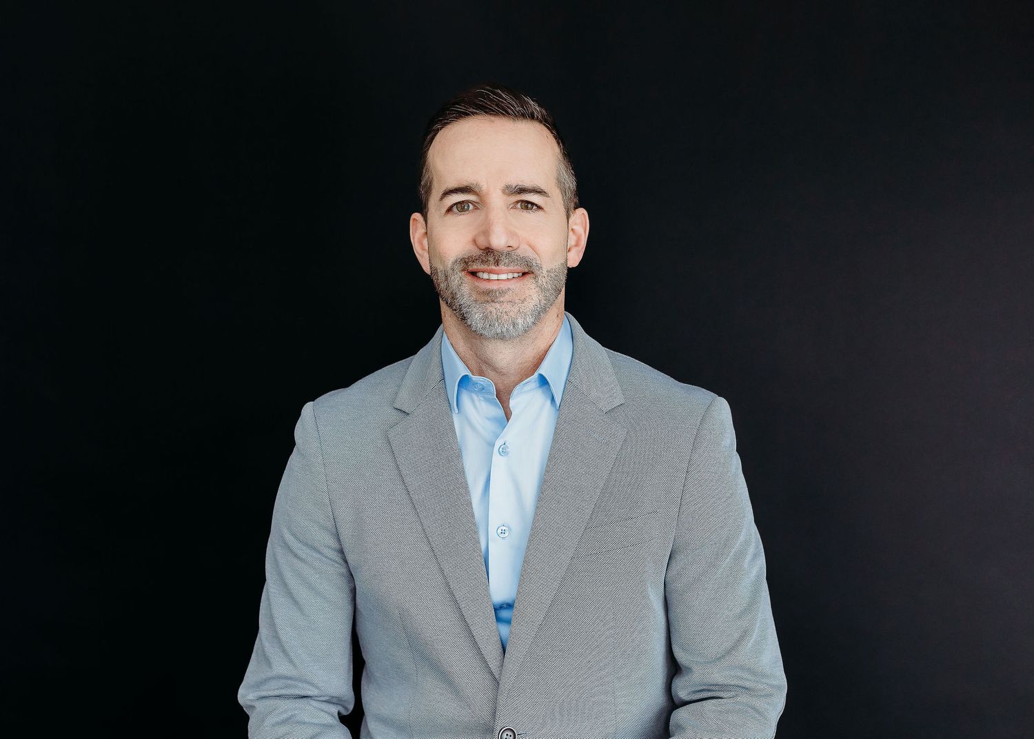 Professional headshot of someone wearing a gray suit jacket and light blue dress shirt against a dark background.