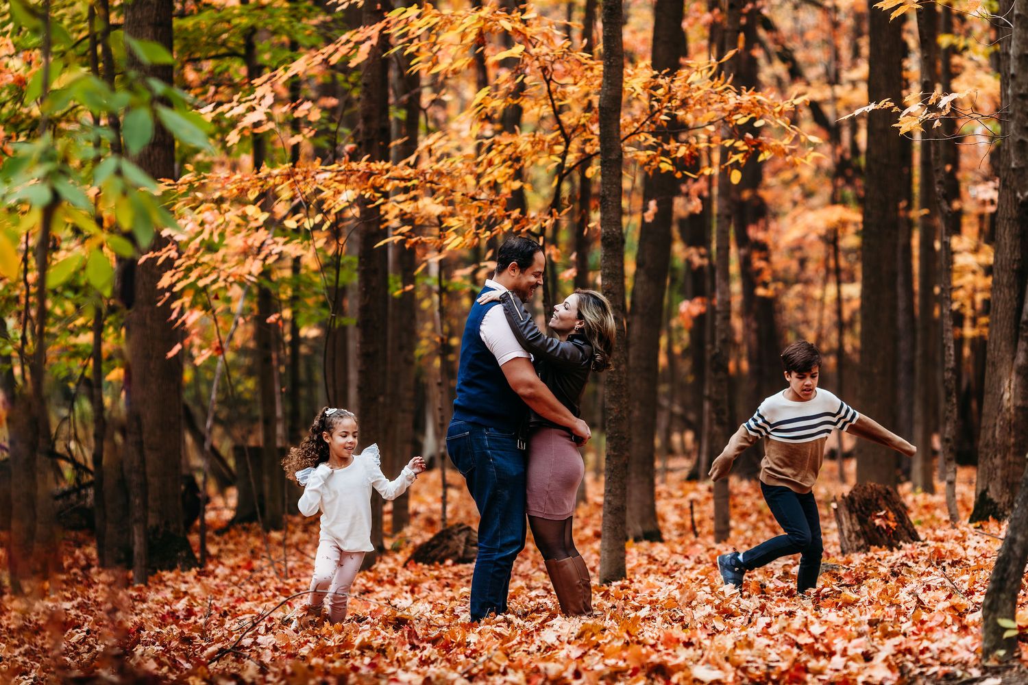 People jump and play in a colorful autumn forest filled with fallen orange leaves and tall trees.