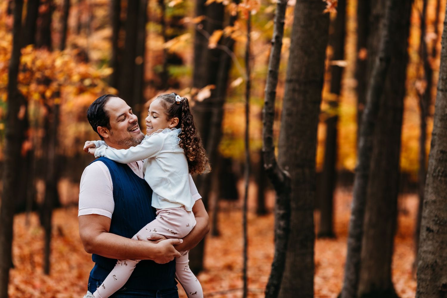 Un parent et son enfant partagent un moment de joie dans une forêt d'automne aux feuilles mortes orangées.