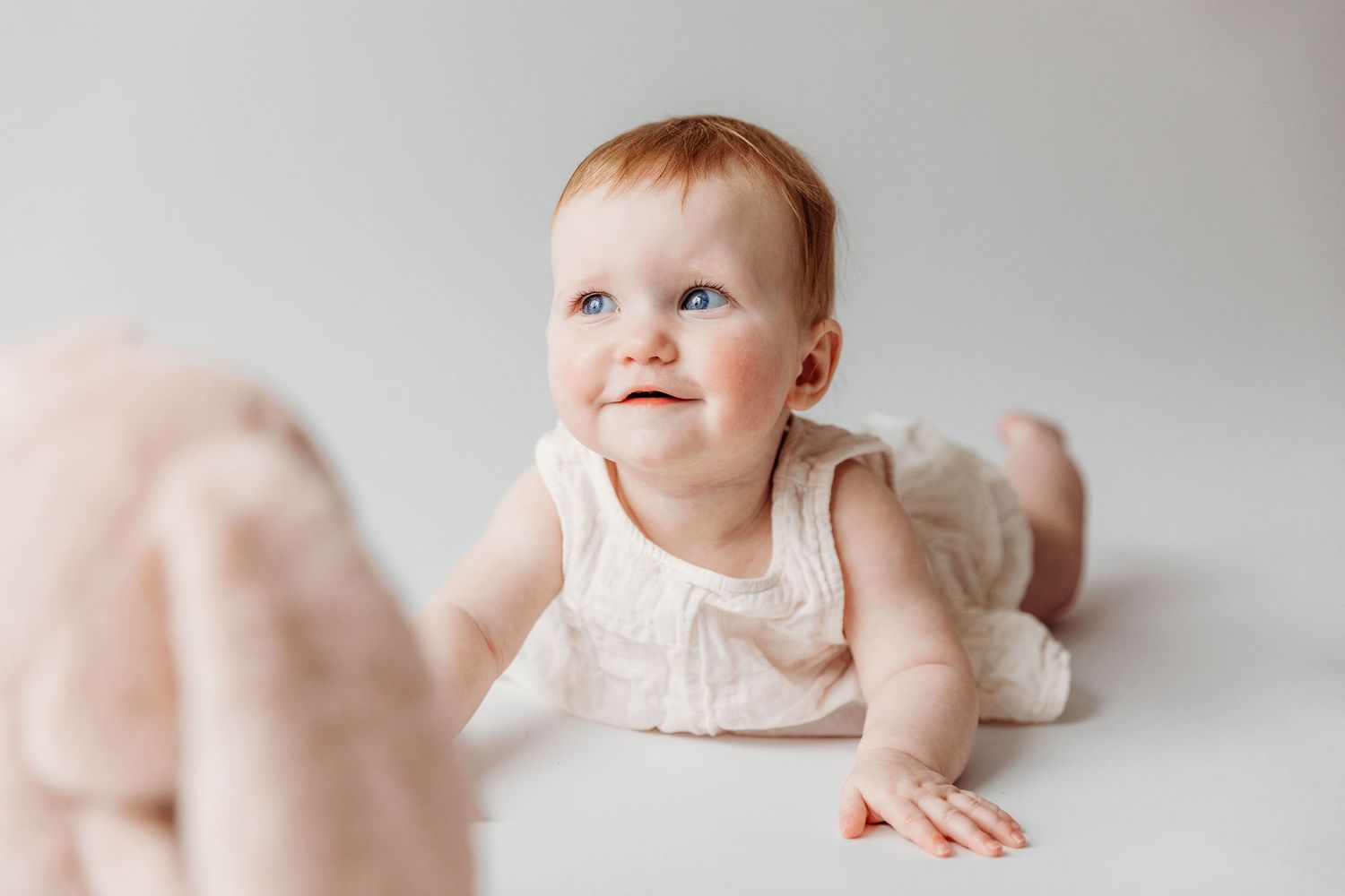 Baby in white sleeveless dress lying on stomach and looking up with a happy expression against light background.