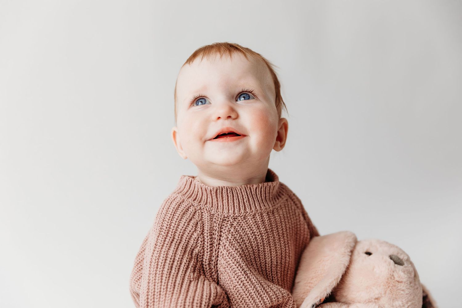 Baby wearing a cozy knit sweater smiles while looking up against a light gray background.