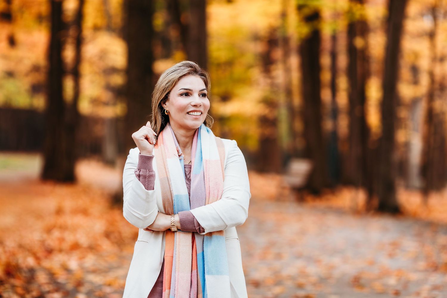 Person wearing a cream sweater and pink scarf poses in a vibrant autumn forest with golden foliage in the background.