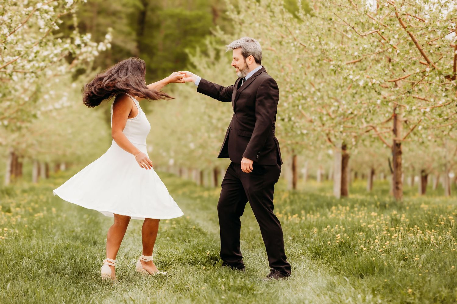 A couple dances playfully together in a lush green orchard on a sunny spring day.