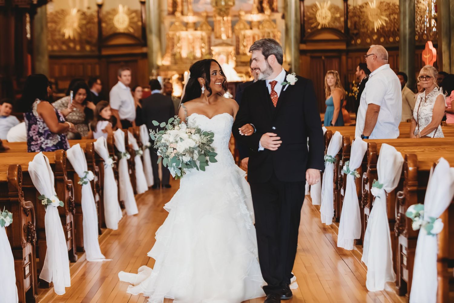 Wedding ceremony guests seated in wooden pews of an ornate church with stained glass windows.