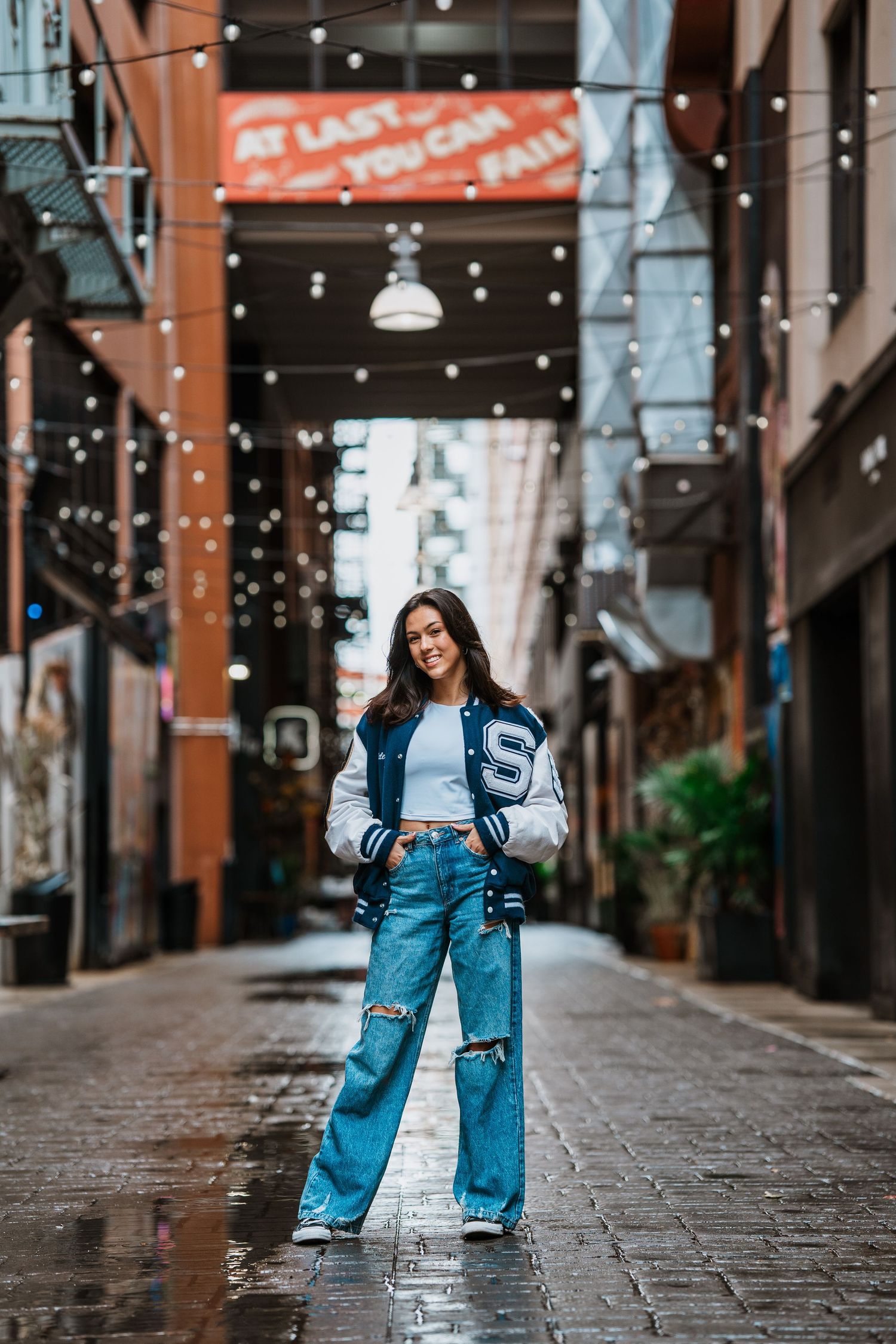 Young woman in varsity jacket and ripped jeans posing confidently in a wet urban alley with string lights.