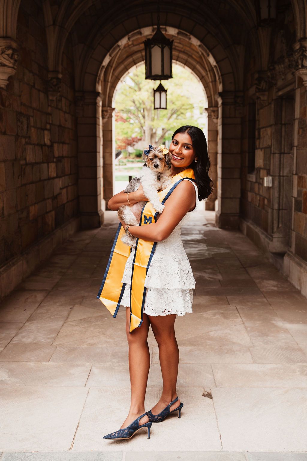 A smiling graduate in a white dress holds a small dog while wearing a gold graduation sash in a stone archway.