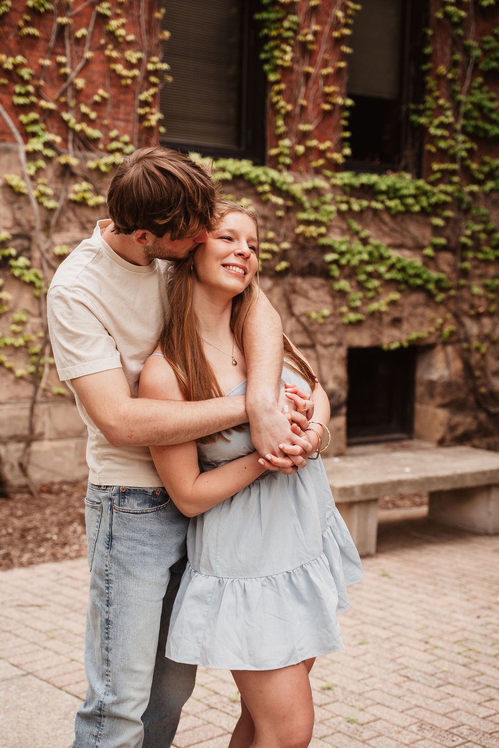 A young couple embraces outdoors, man kissing woman's cheek as she smiles, ivy-covered brick wall behind them.