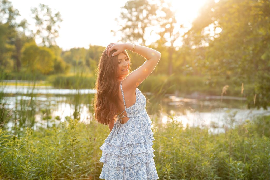 Woman in floral dress stands in golden sunlight by lake during sunset with trees in background.