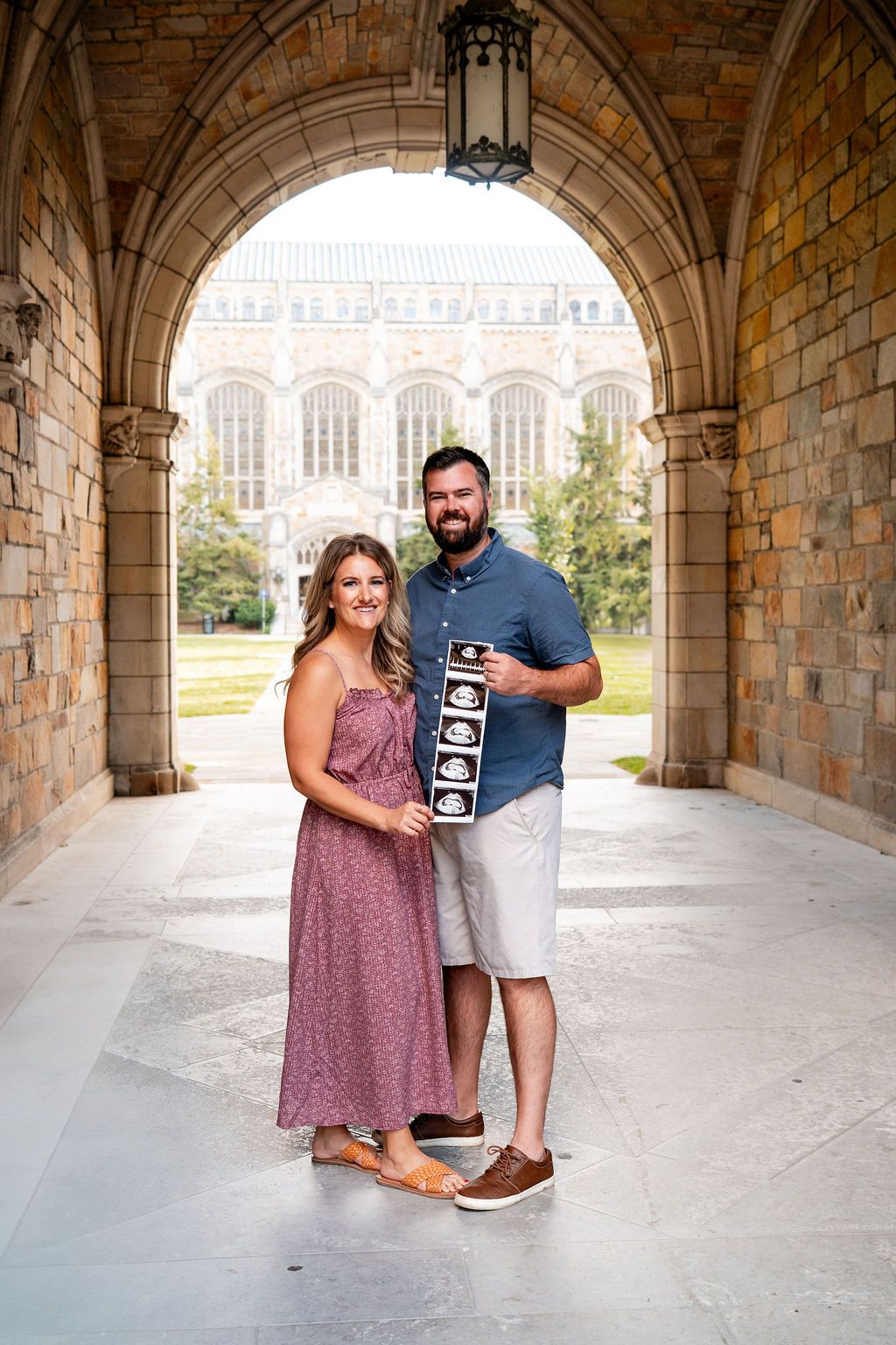 A couple stands under a stone archway holding ultrasound photos and smiling together in a romantic pregnancy announcement.