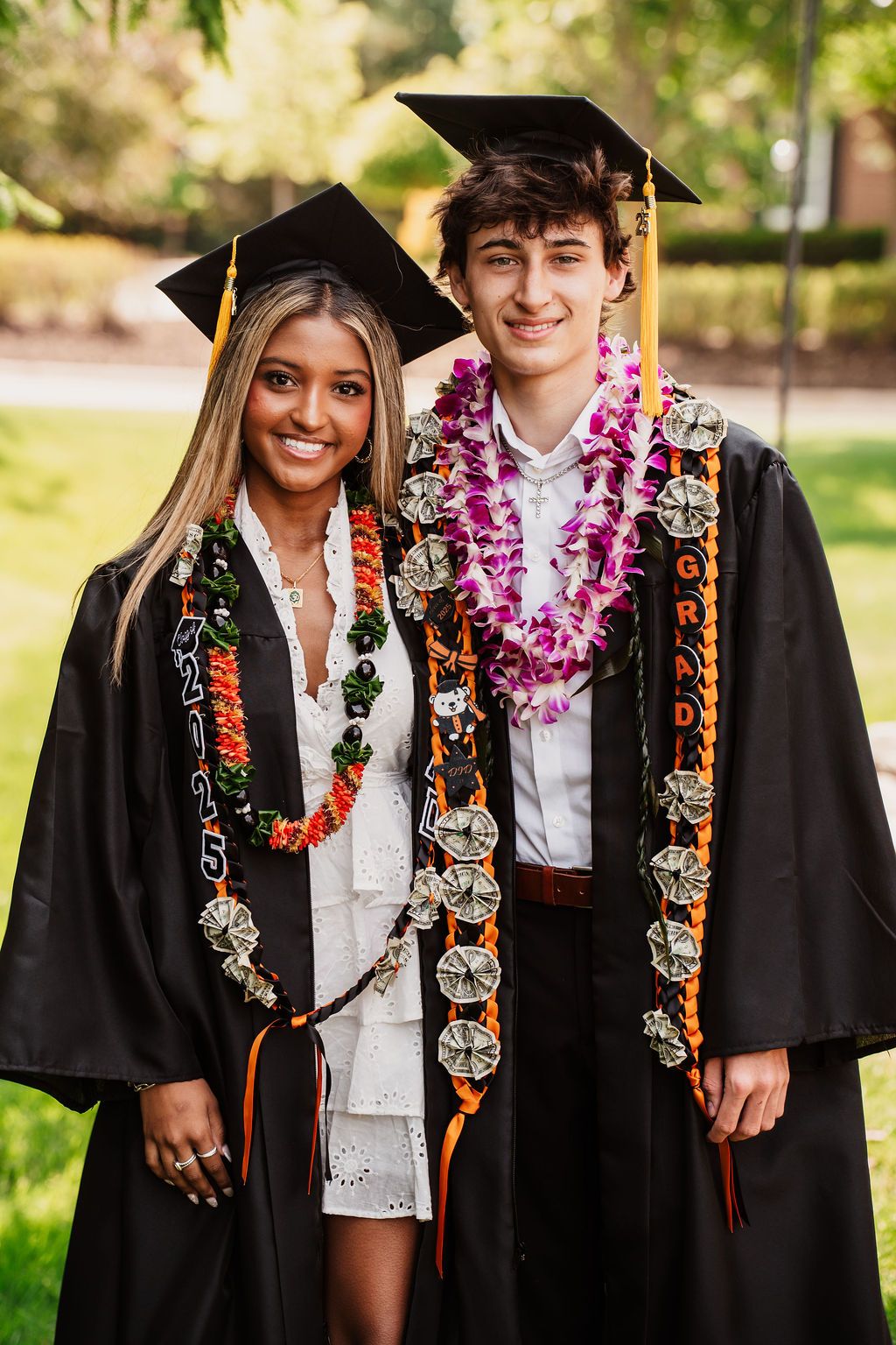 Graduates wearing black caps and gowns with colorful leis and decorative sashes celebrate outdoors on a sunny day.