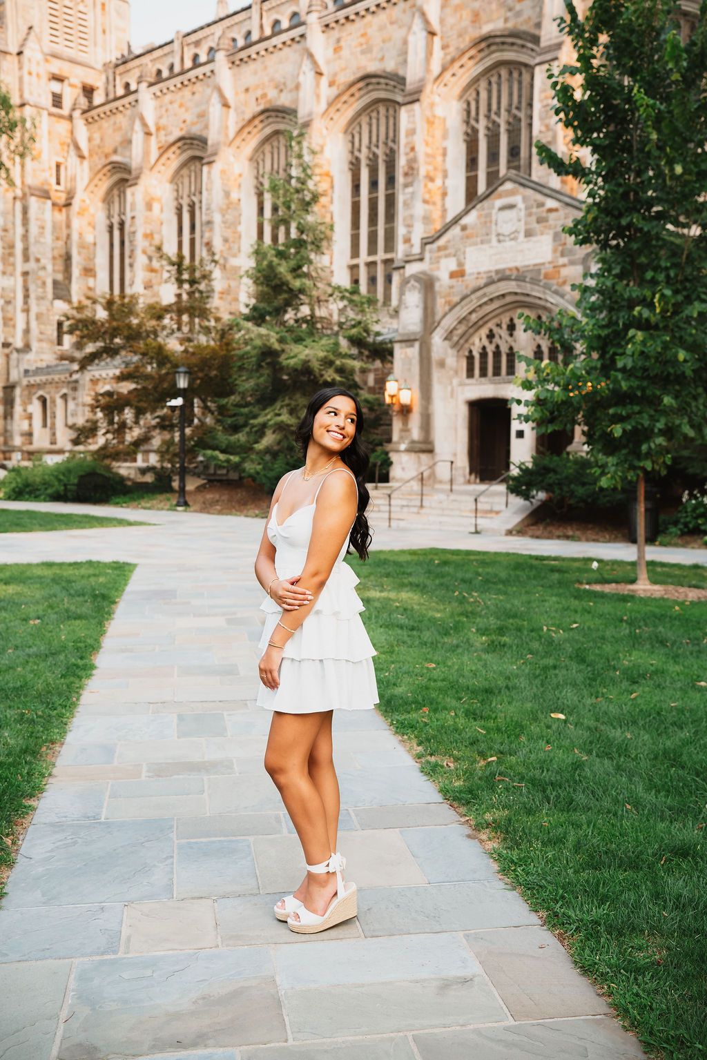 A person in a white dress poses for graduation photos in front of a Gothic university building.