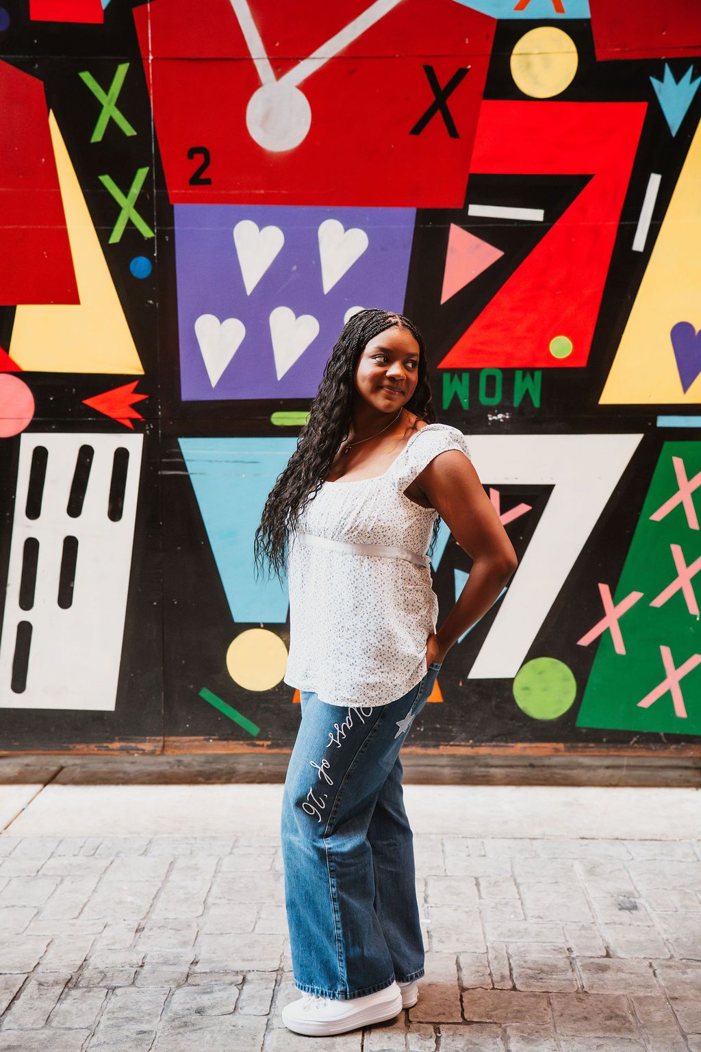 Woman in white top and jeans poses against colorful geometric mural wall with hearts and shapes.