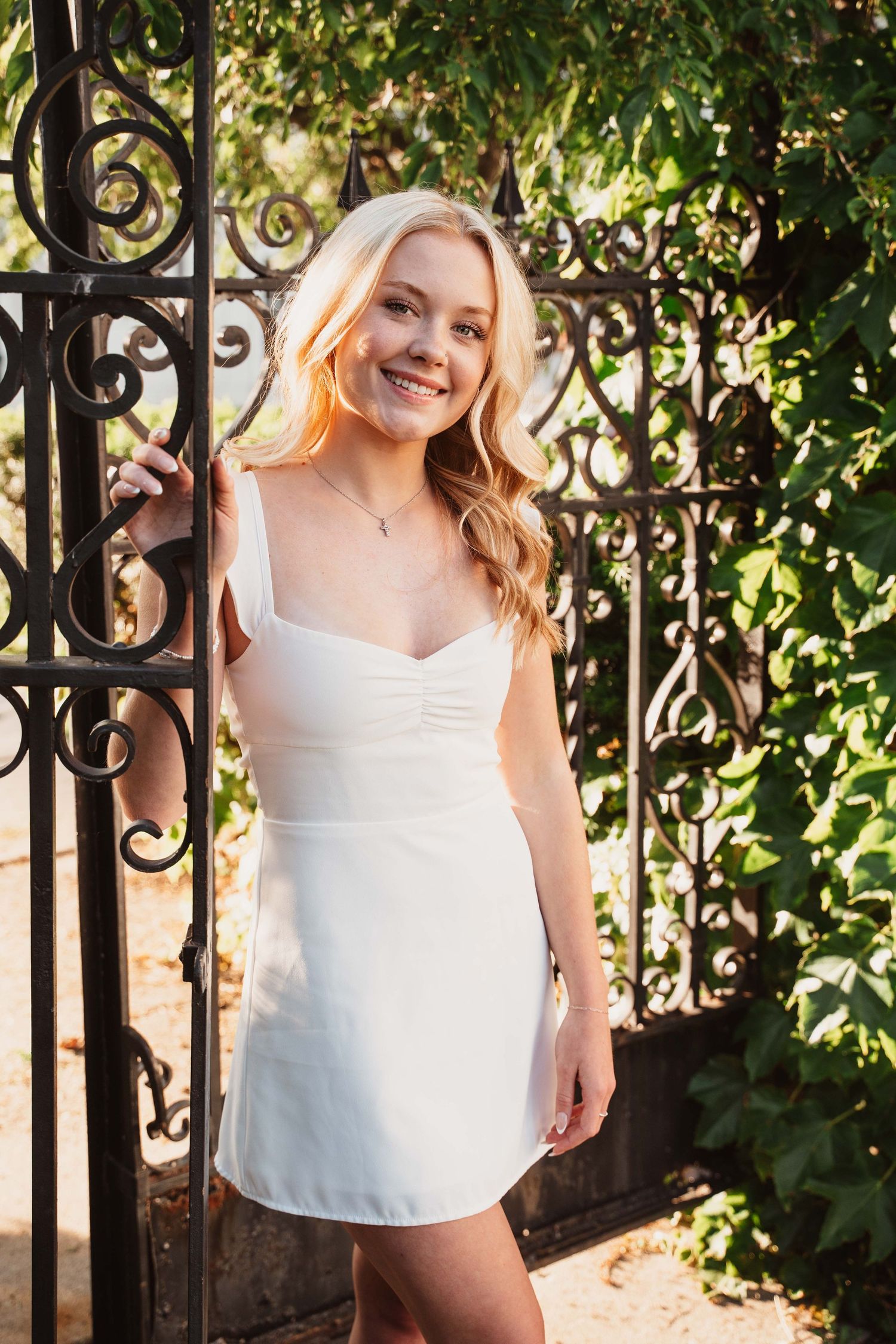 Smiling blonde woman in white mini dress posing by ornate iron gate surrounded by lush green ivy.