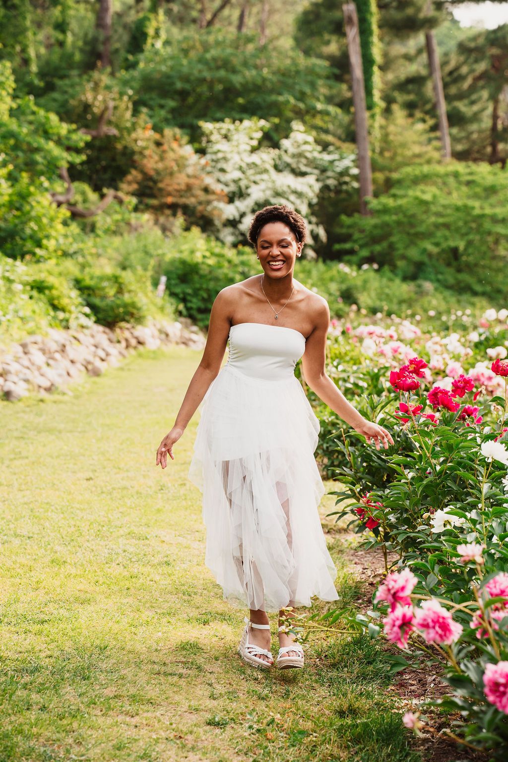 A person in a flowing white dress dances joyfully among blooming pink roses in a sunlit garden.