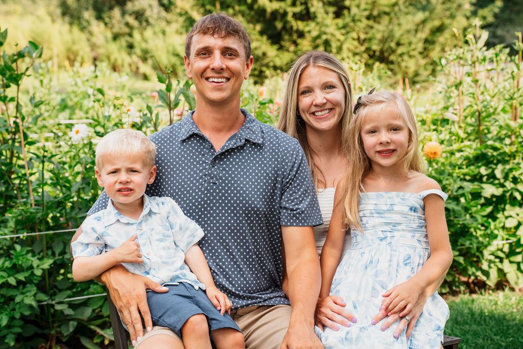 Happy family of four poses outdoors in a lush green garden, parents smiling with young boy and girl.