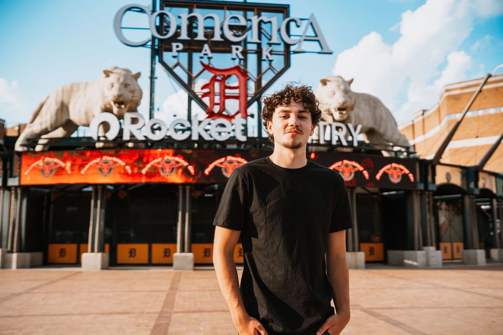 Young man in black t-shirt stands before Comerica Park entrance with tiger statues in Detroit, Michigan.