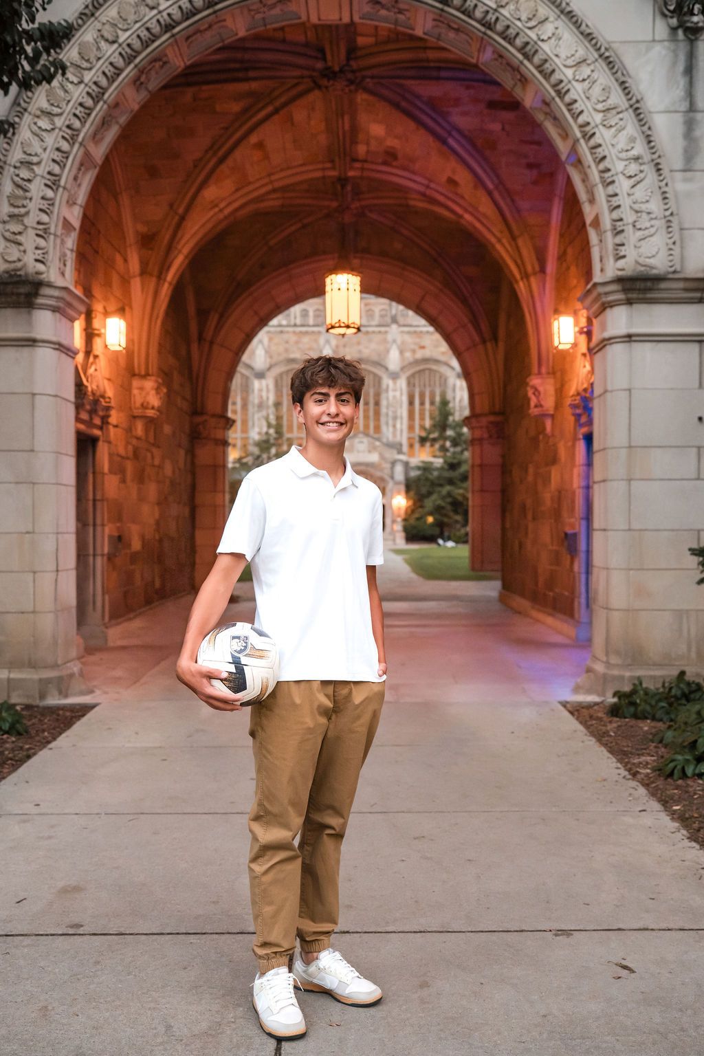 Young man holding a soccer ball stands smiling under a Gothic stone archway on a college campus at dusk.