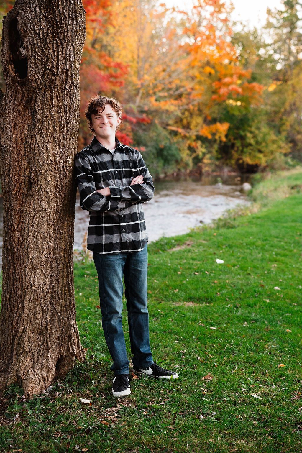 Teen boy in plaid shirt leaning against tree with arms crossed, smiling outdoors in autumn park setting.