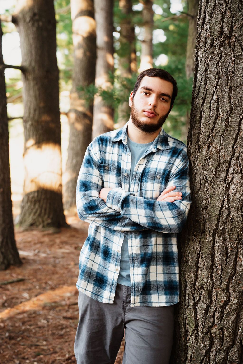 Young man with beard in plaid shirt leaning against tree trunk in forest with arms crossed.