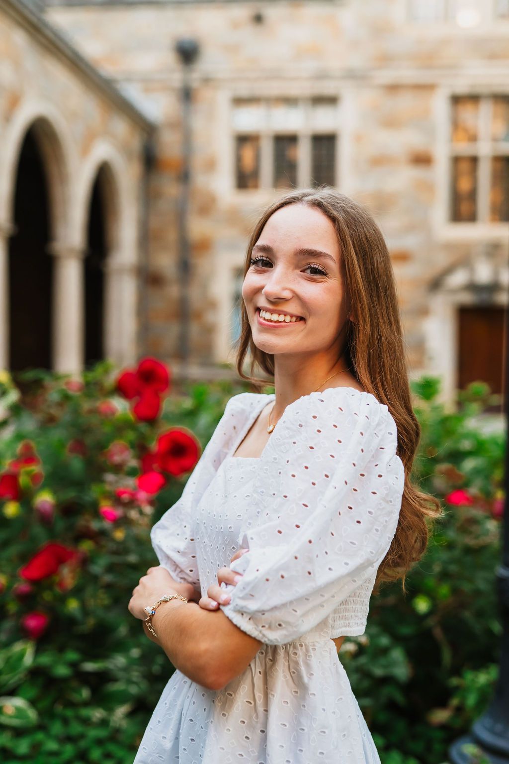 A person in a white eyelet dress smiles while standing in front of red roses and stone archways.