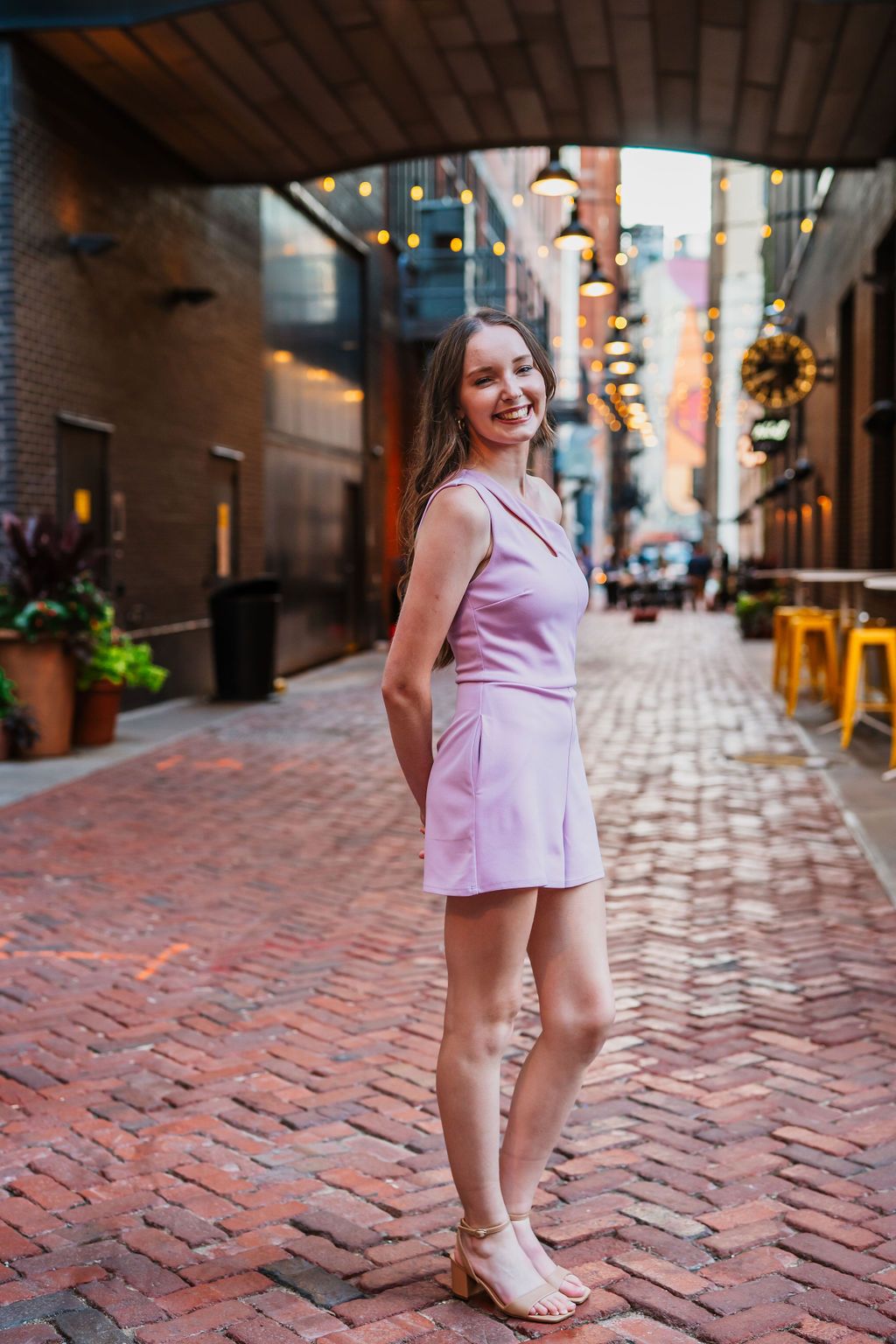 Young woman in pink mini dress smiling on charming brick alley with string lights in urban setting.