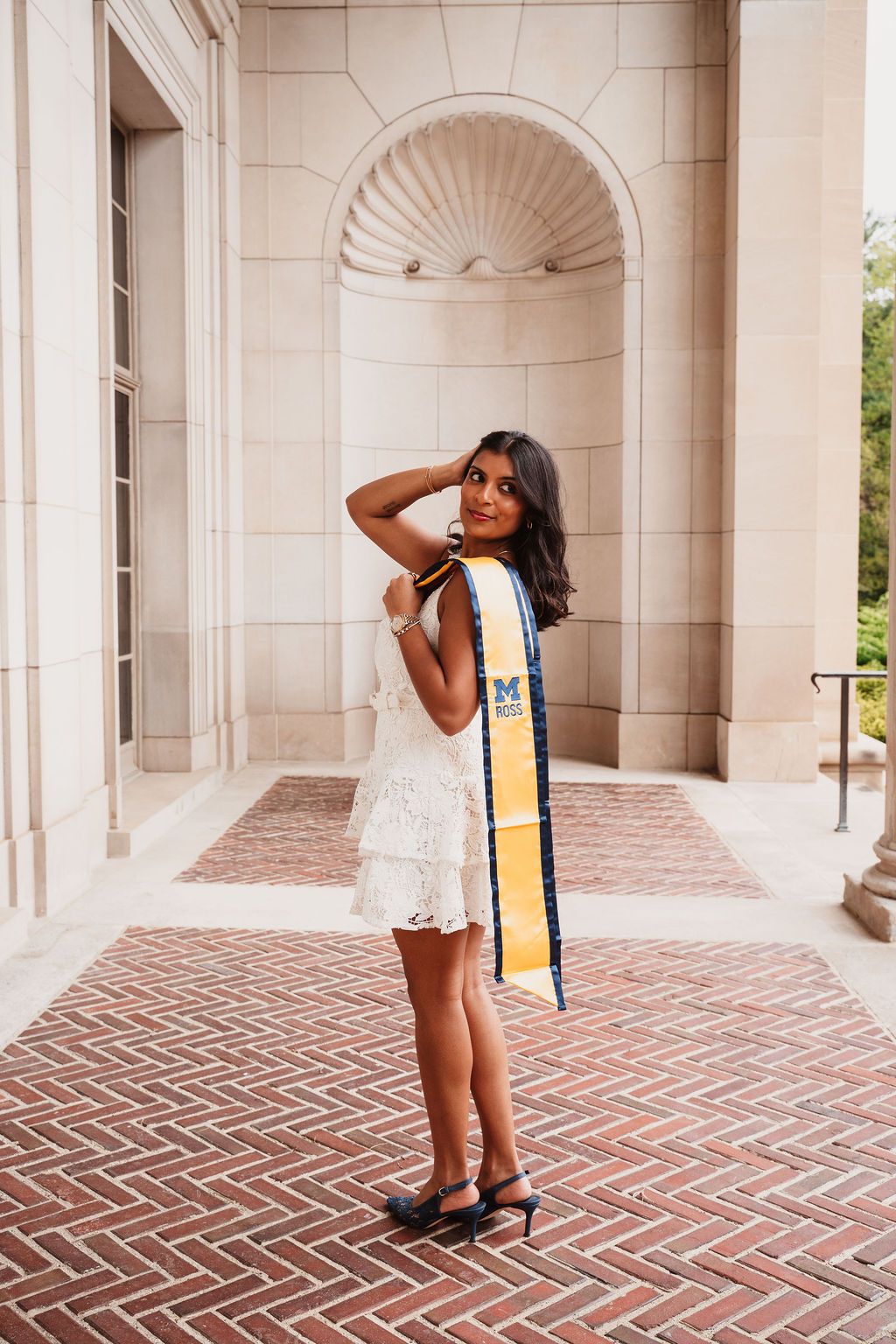 Young woman in white lace dress with University of Michigan Ross graduation sash poses outdoors on brick walkway.