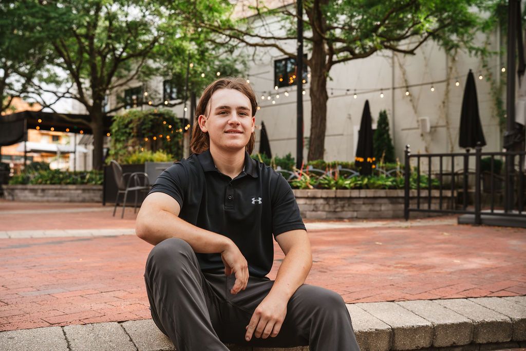 Young man in black Under Armour polo sits on brick steps in an outdoor urban plaza with string lights.