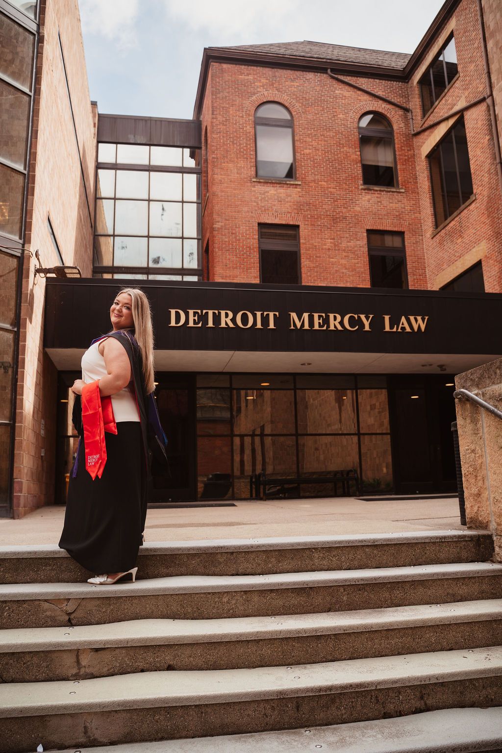 Graduate poses in cap and gown regalia outside Detroit Mercy Law School building entrance on graduation day.