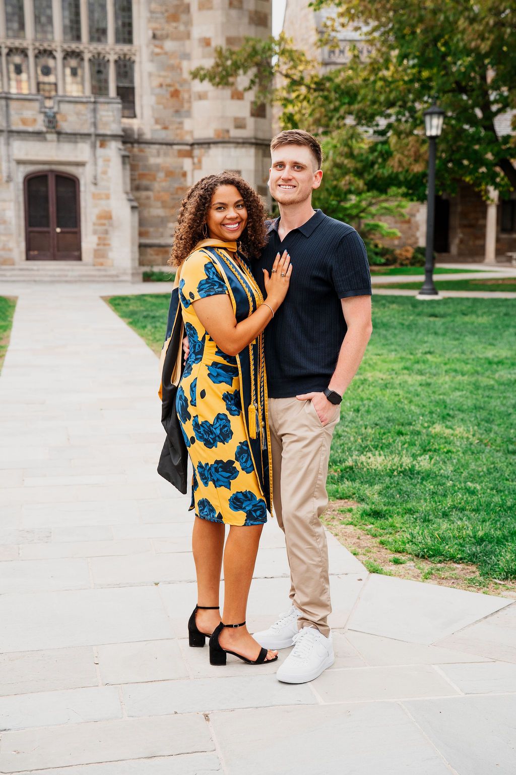 A couple poses together in front of a collegiate Gothic building on a sunny day.