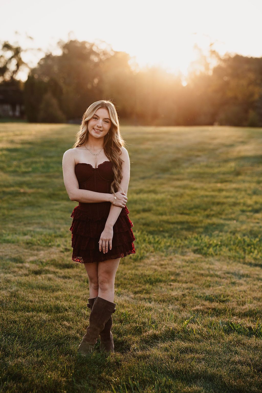 Young woman in dark red strapless dress and brown boots poses in sunlit green field at golden hour.